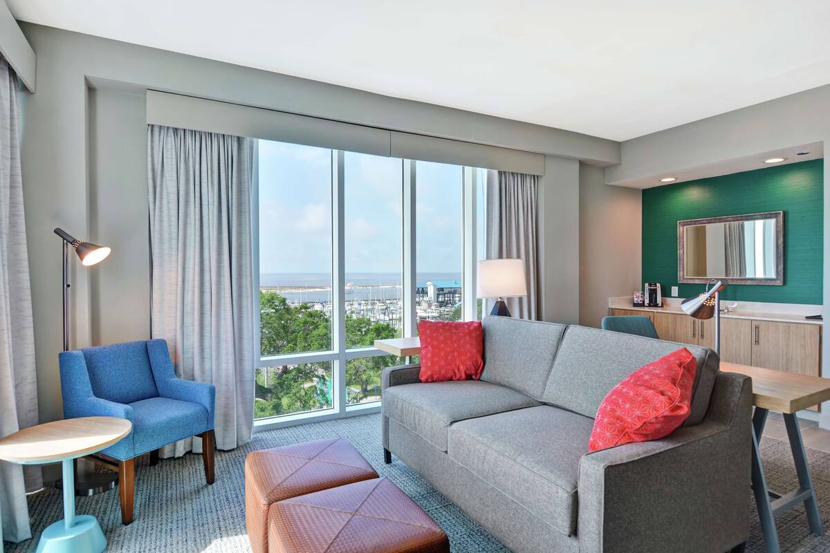 A modern hotel room with a large window, a grey sofa, a blue armchair, and a small round table.