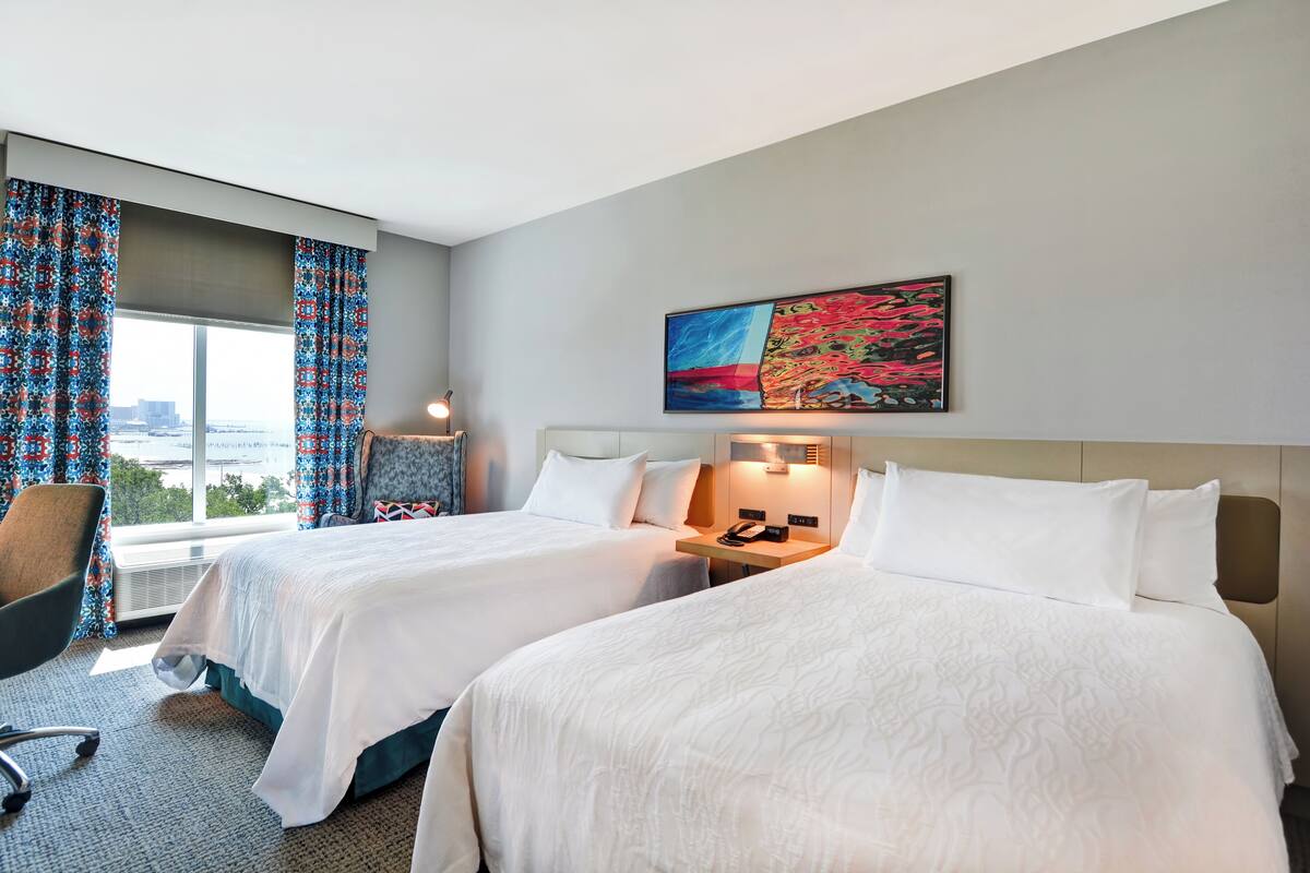A hotel room with two beds, a desk, a chair, and a large window with curtains.