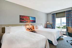 A hotel room with two beds, a painting, a desk with a phone, a chair, and a large window with curtains. - Hilton Garden Inn Biloxi (Biloxi)