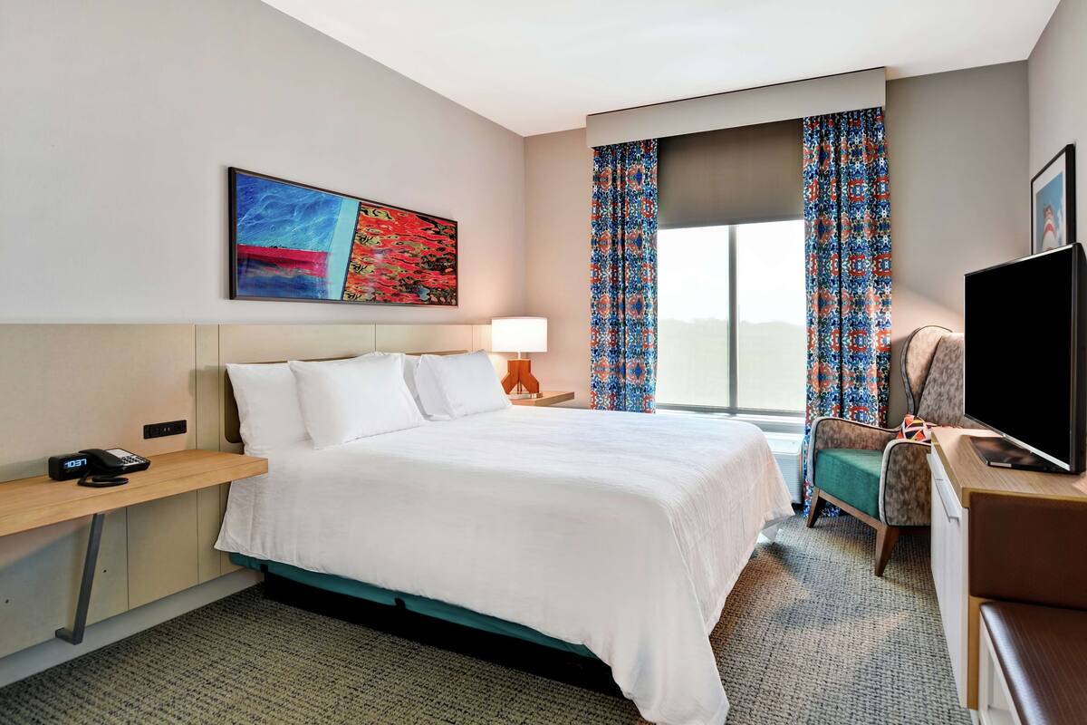 A hotel room with a bed, a desk with a lamp, a television, and a window with patterned curtains.