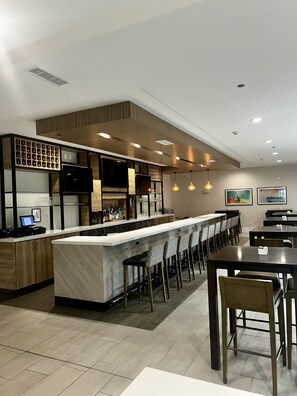 A modern bar area with a long counter, bar stools, and a seating area with tables and chairs. - Hilton Garden Inn Biloxi (Biloxi)