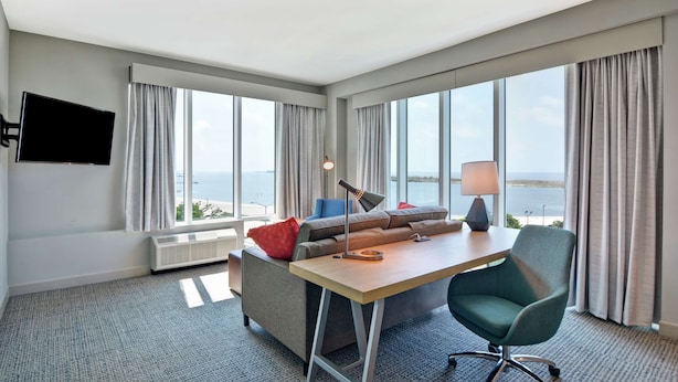 A hotel room with a large window, a desk, a chair, a sofa, and a TV. - Hilton Garden Inn Biloxi (Biloxi)