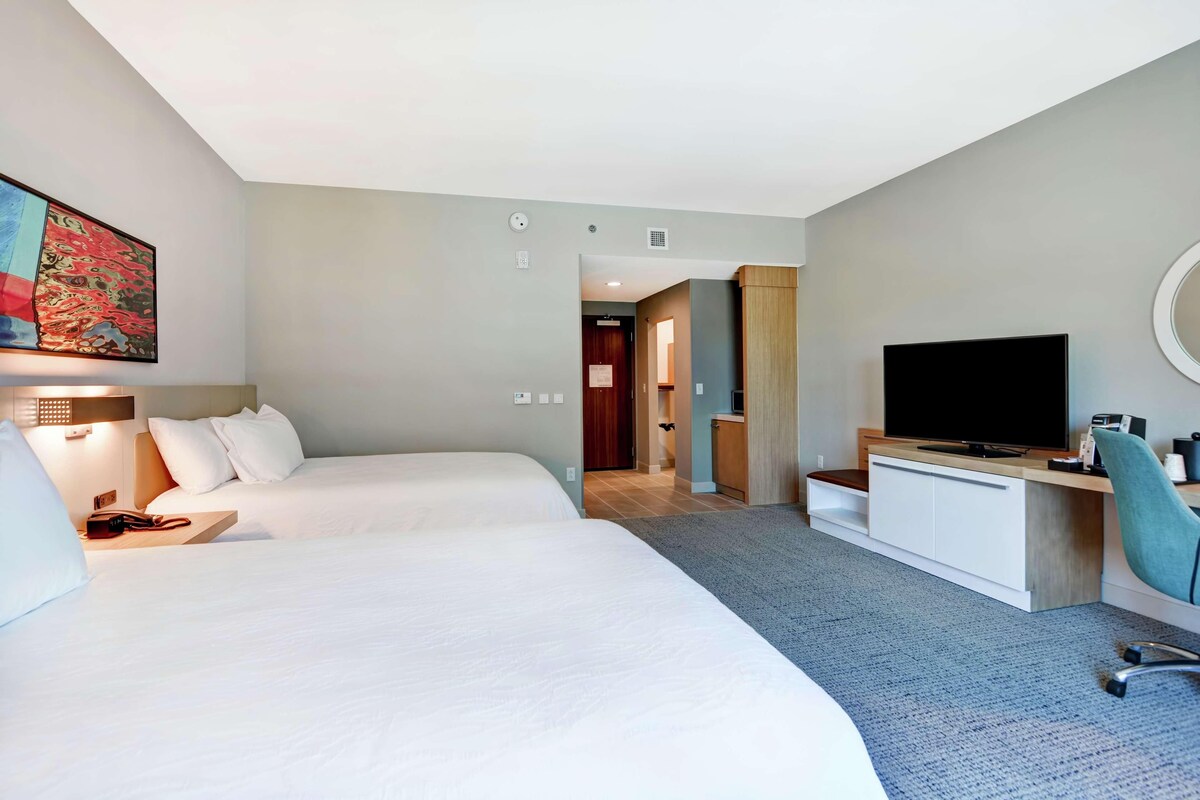 Habitación de hotel con dos camas, televisión, escritorio con silla y un cuadro en la pared.