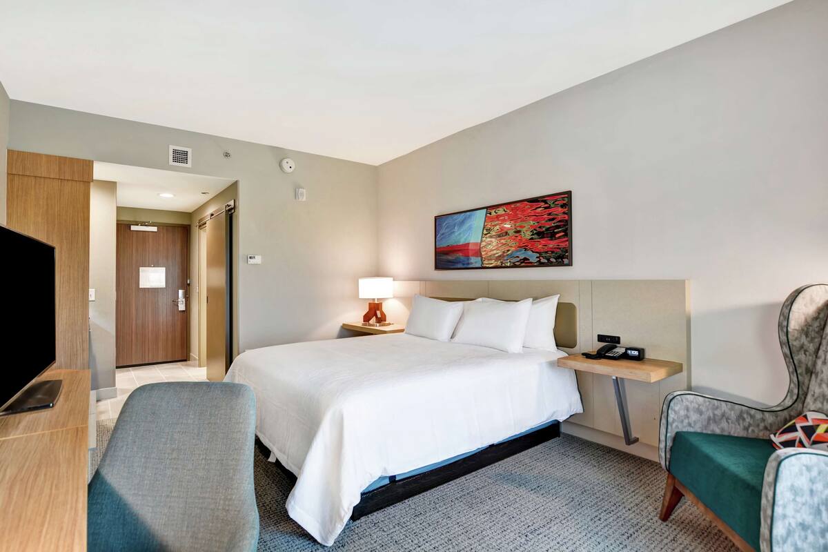 A hotel room with a bed, a desk, a chair, a TV, a painting on the wall, and a door leading to another room.