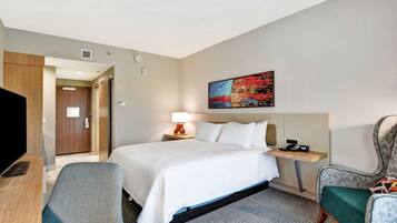 A hotel room with a bed, a desk, a chair, a TV, a painting on the wall, and a door leading to another room.