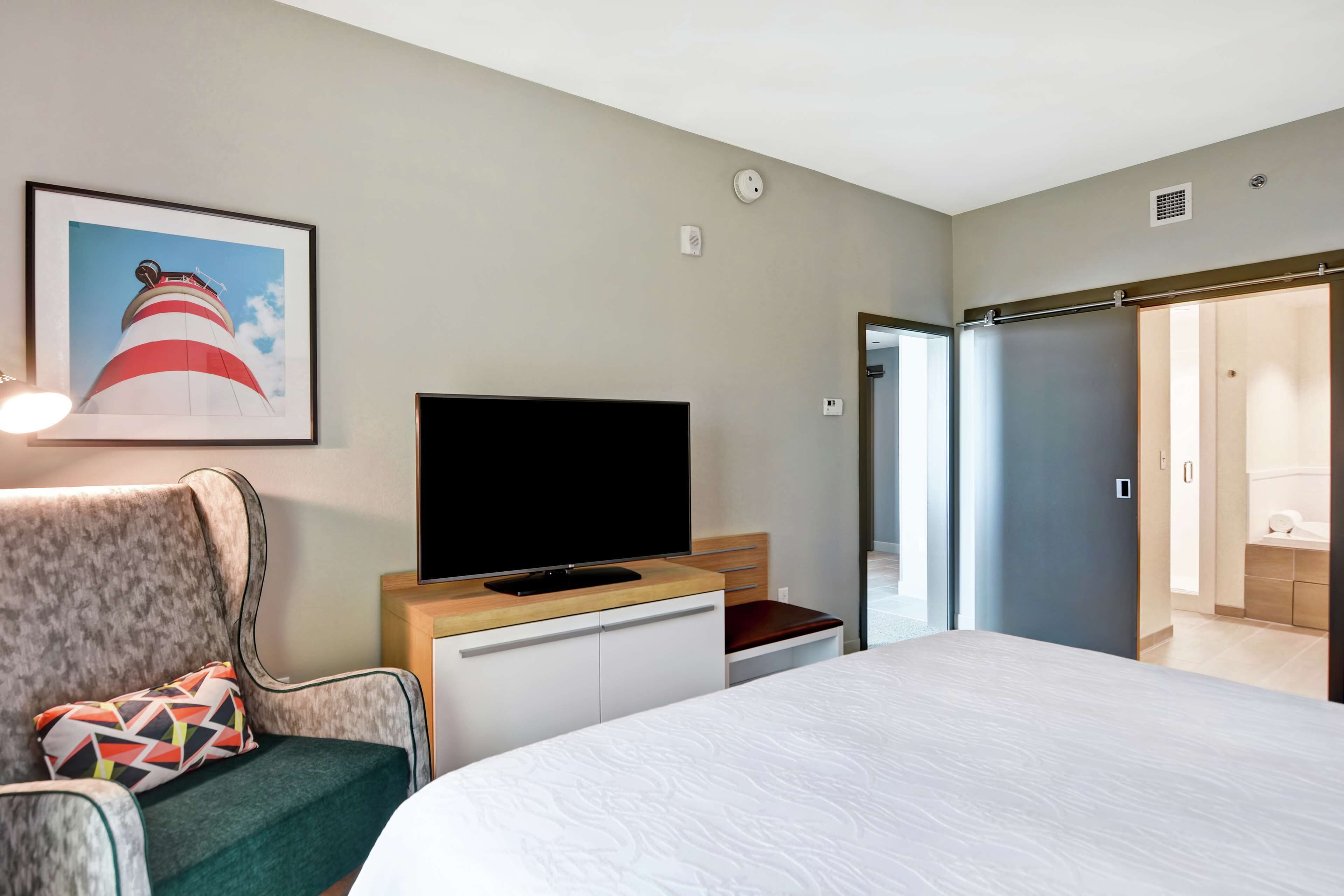 Une chambre d’hôtel avec un lit, un téléviseur posé sur un meuble en bois, une chaise avec un coussin coloré, un tableau encadré représentant un phare et une armoire avec une porte coulissante.