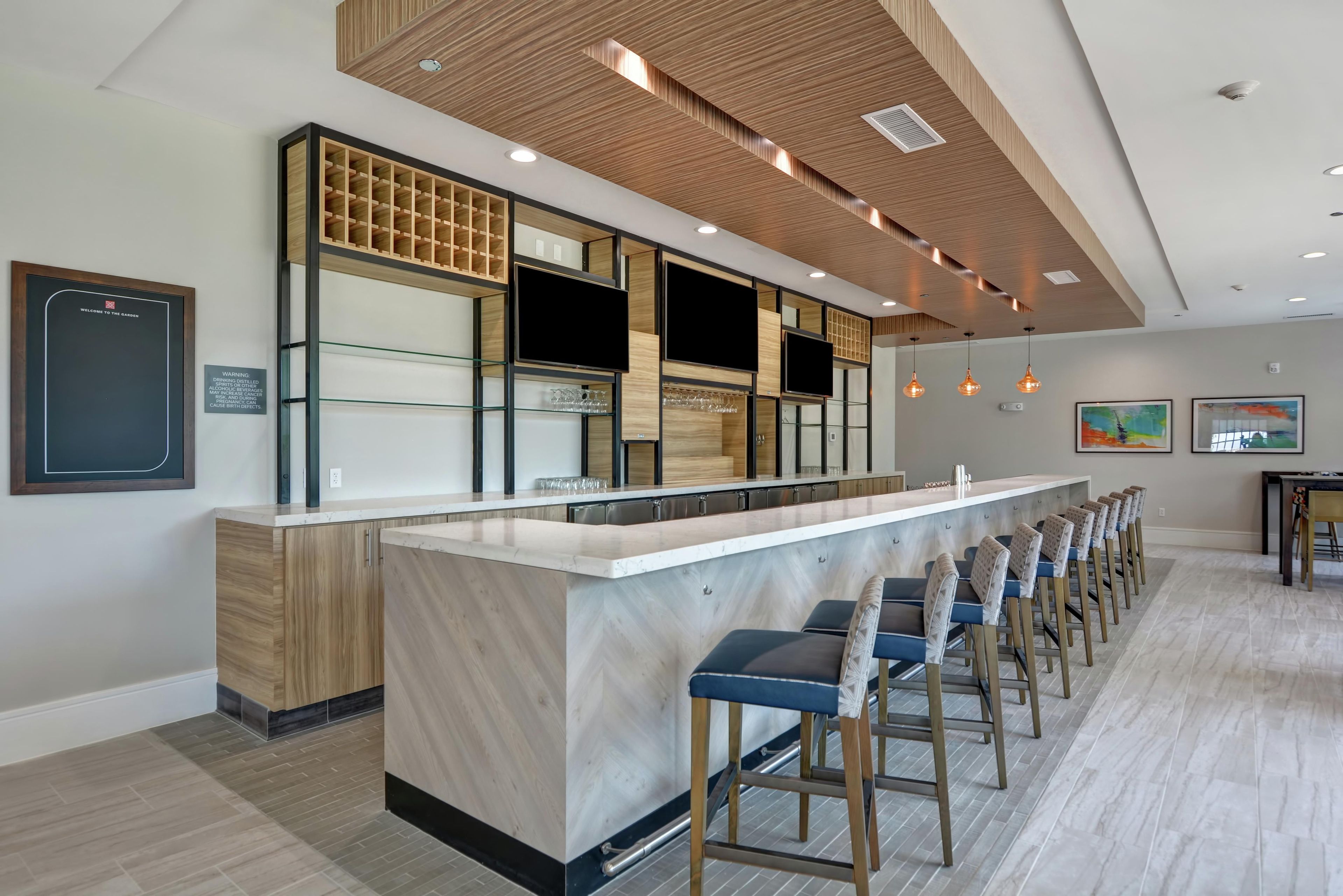 A modern bar area with a long counter and multiple stools, a wooden ceiling, and a wall-mounted TV.