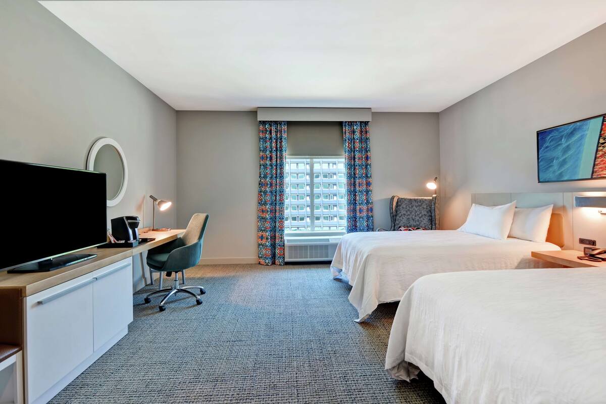 A hotel room with two beds, a desk with a computer, a TV, and a window with patterned curtains.
