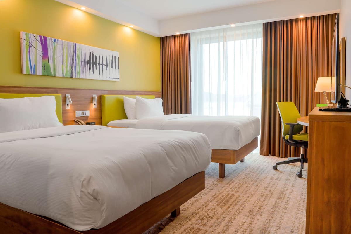 Quarto de hotel com duas camas, uma escrivaninha com cadeira, um quadro na parede e uma janela grande com cortinas.
