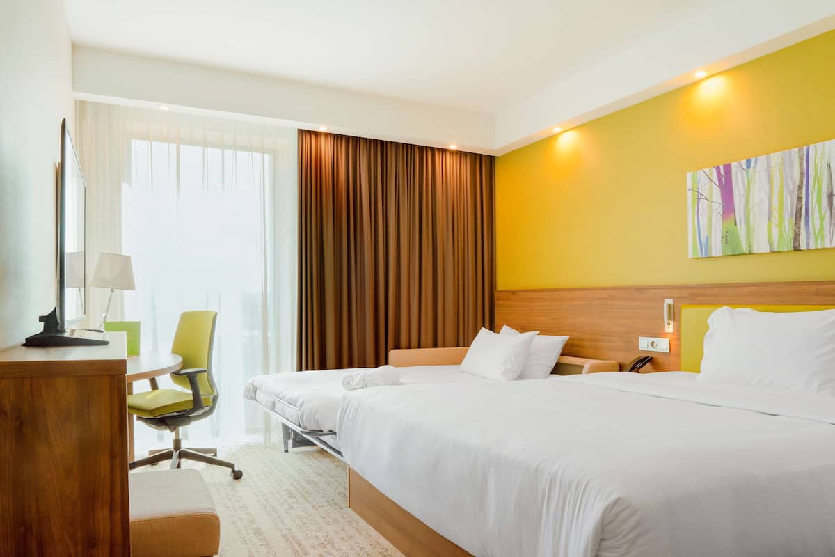 Quarto de hotel com duas camas, uma escrivaninha com luminária, uma cadeira e uma janela grande com cortinas.