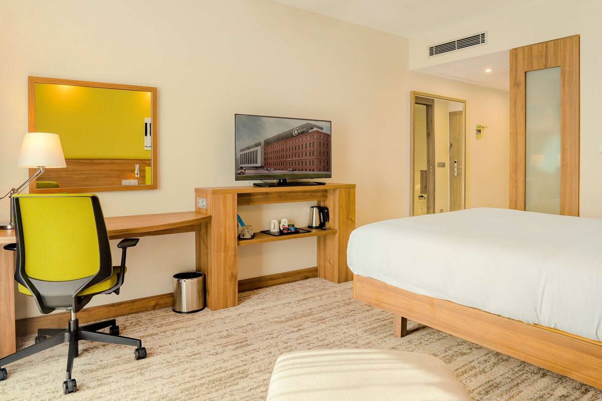 Quarto de hotel com cama, escrivaninha com cadeira, televisão, espelho e prateleira com cafeteira.