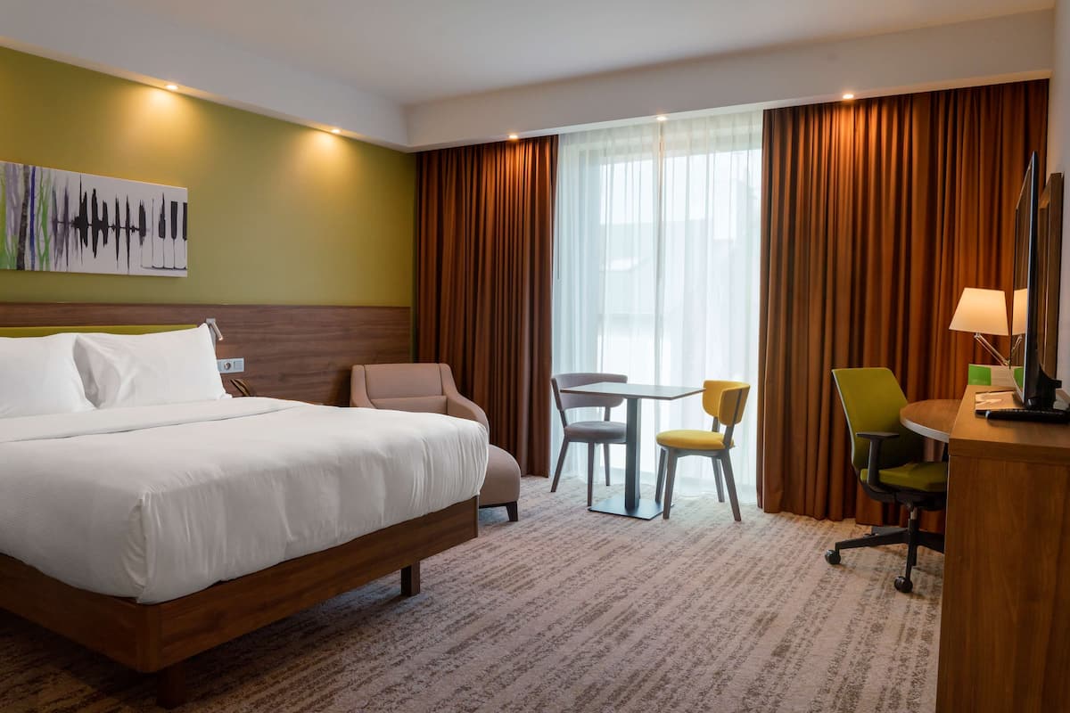 Quarto de hotel com uma cama grande, uma escrivaninha com cadeira, uma mesinha com duas cadeiras e uma janela ampla com cortinas.