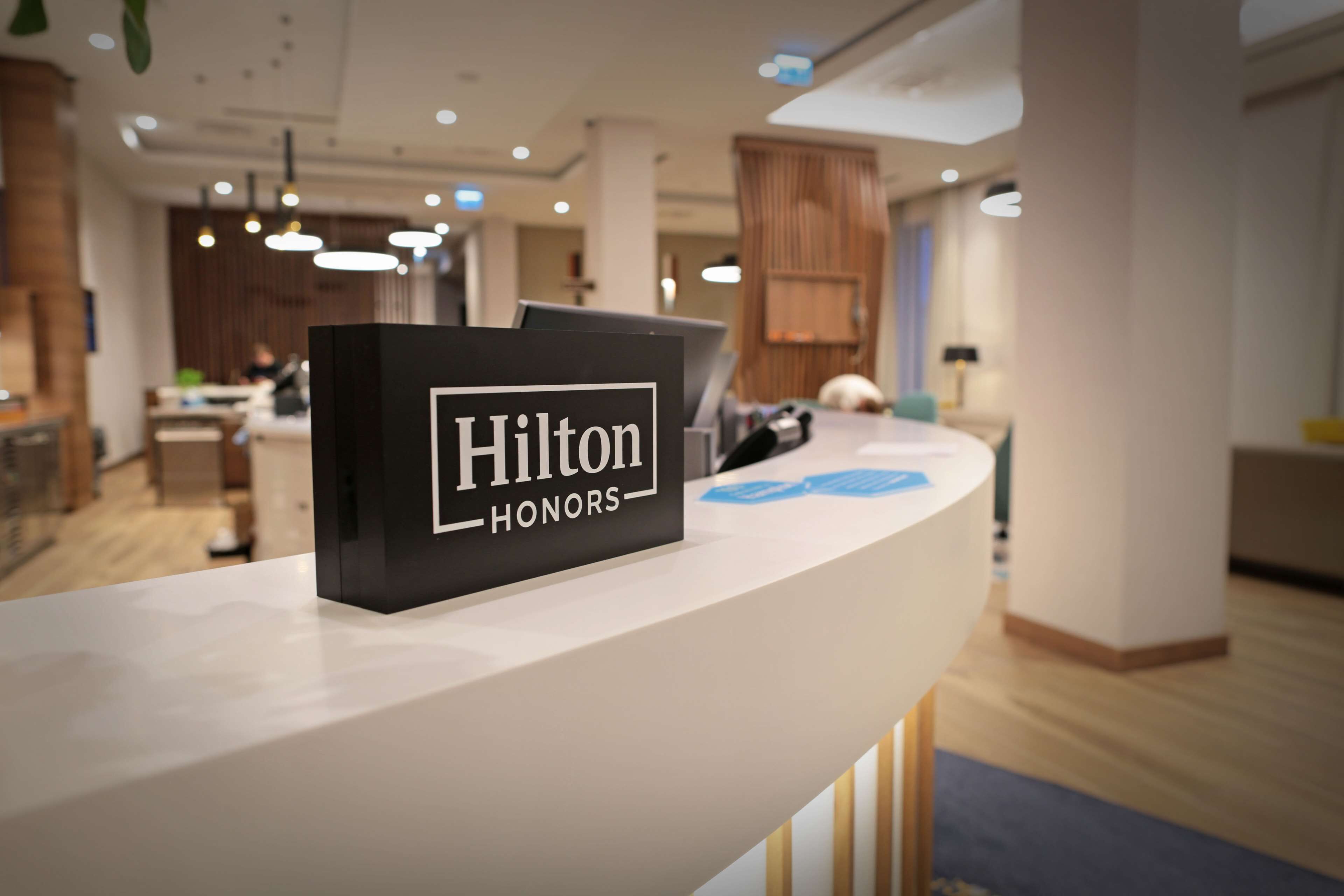 A reception desk with a 'Hilton Honors' sign, a modern interior with wooden accents, and a visible dining area in the background.