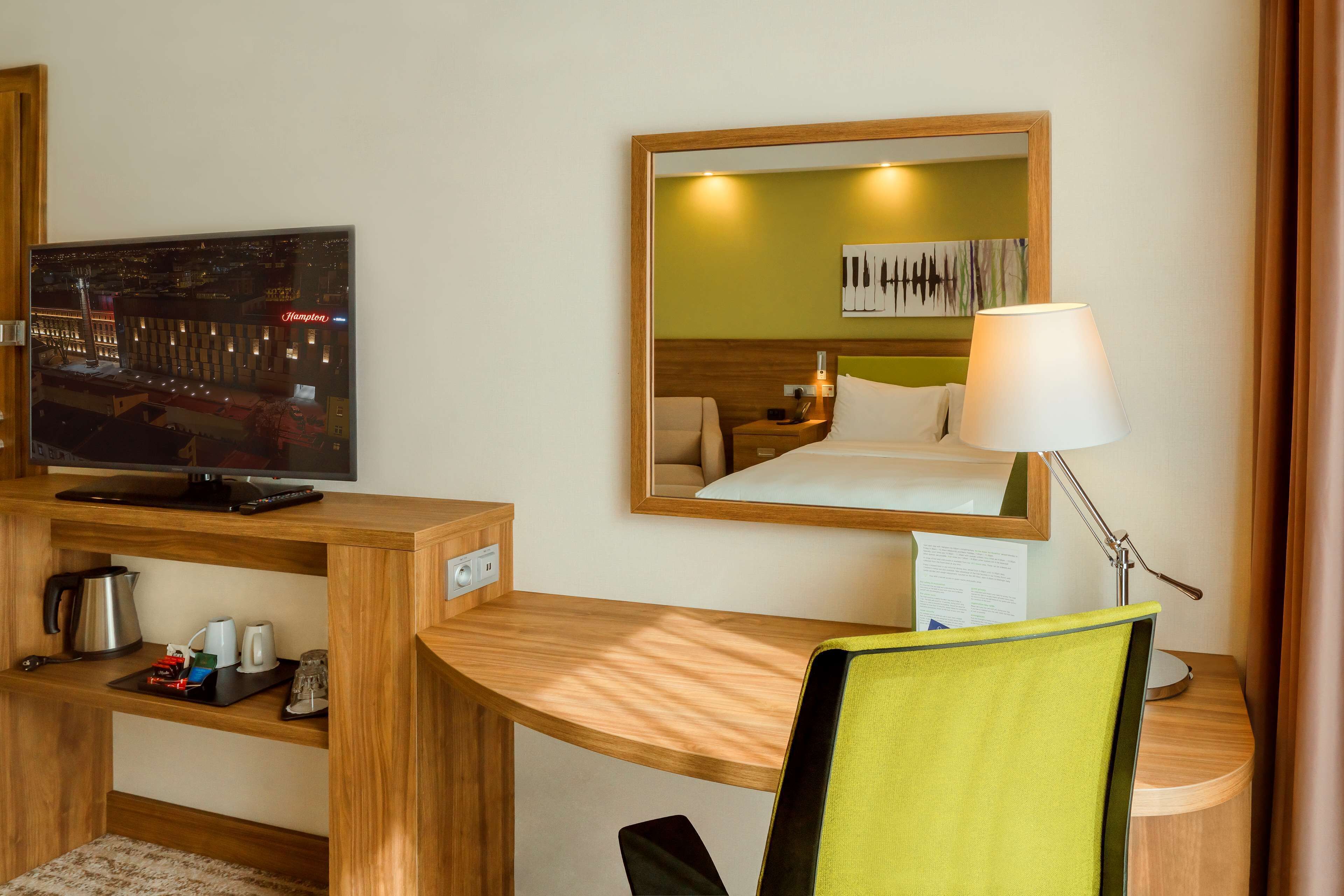 A hotel room with a wooden desk, a television, a bed with a green headboard, a mirror, and a lamp.