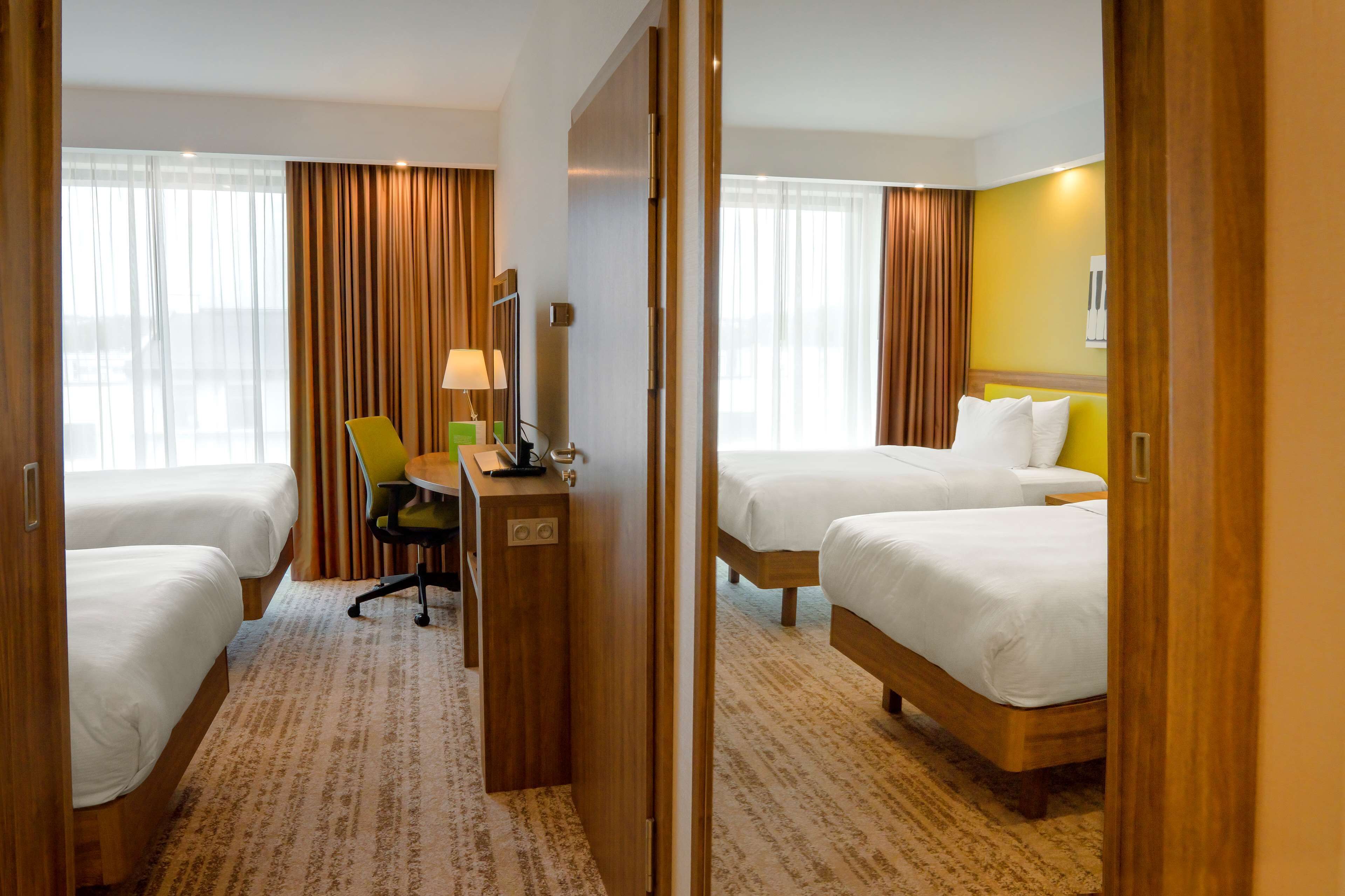 Habitación de hotel con dos camas individuales, un escritorio con silla, una lámpara y un ventanal con cortinas.