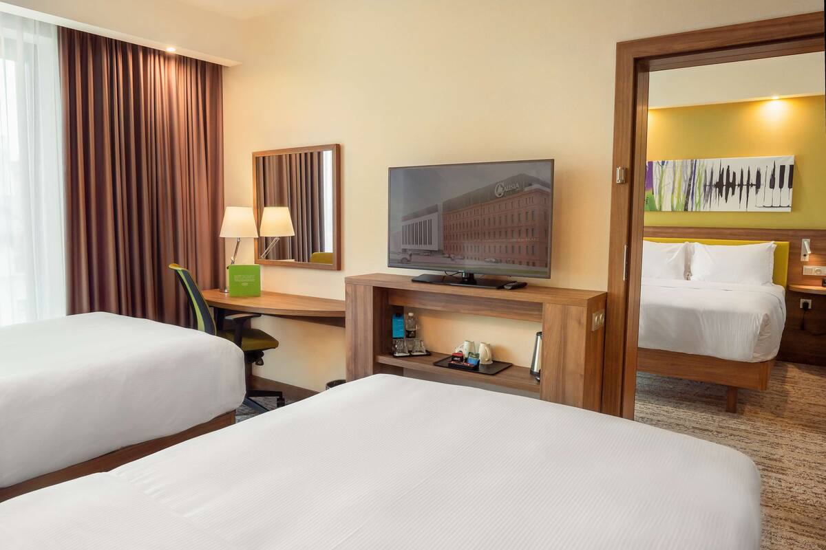 Quarto de hotel com duas camas, televisão, escrivaninha com luminária e espelho.