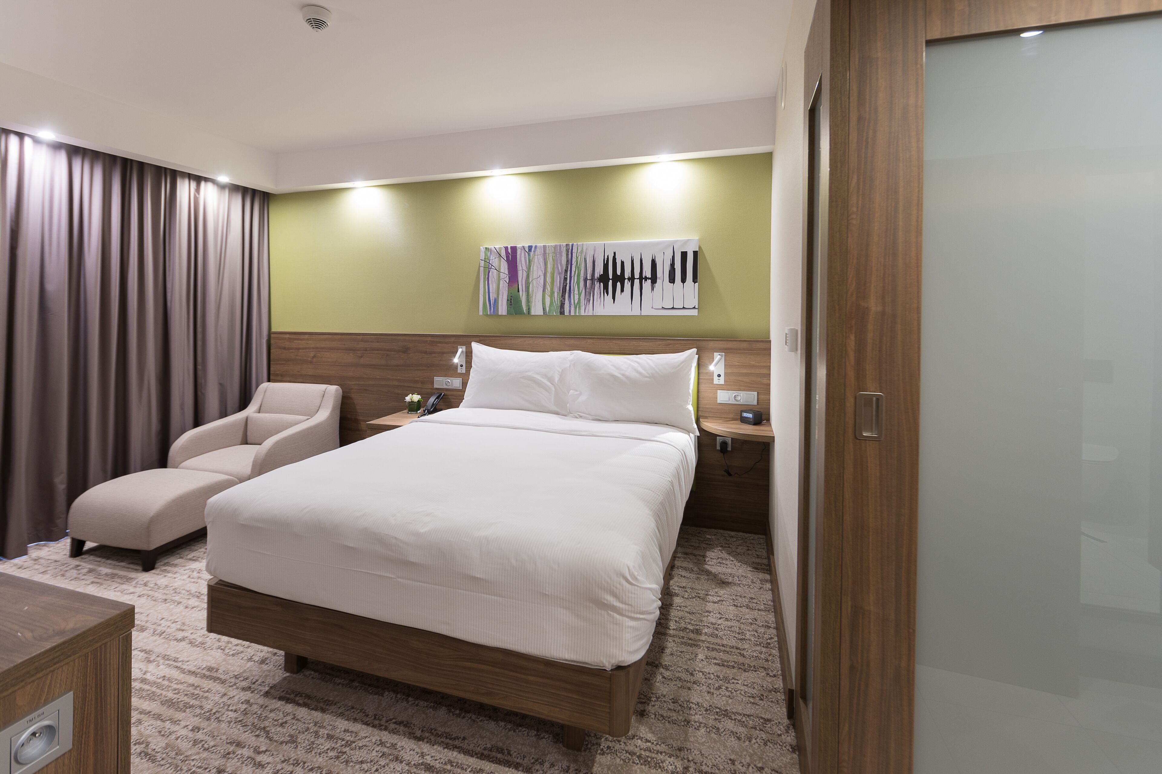 A modern hotel room with a large bed, a chair, a bedside table, and a wall-mounted artwork.