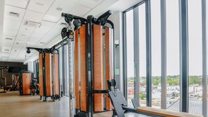 A gym with exercise equipment and a bench, large windows, and a view of the city. - Hampton by Hilton Kalisz (Kalisz)