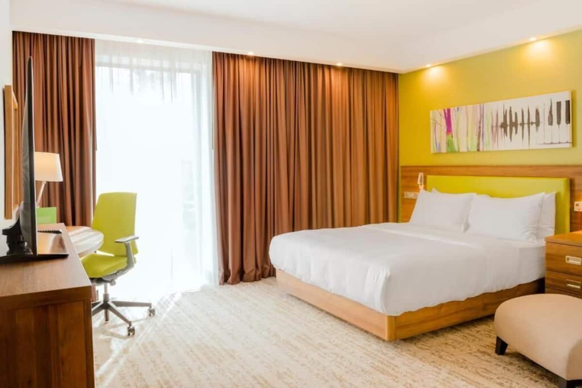 Quarto de hotel com cama, escrivaninha, cadeira, luminária e um quadro na parede.