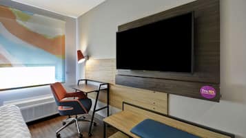 Una habitación de hotel moderna con una televisión de pantalla plana grande montada en la pared, un escritorio con una silla, un banco y una cama con cabecero estampado.