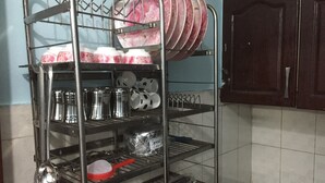 Un étagère en métal contenant des ustensiles de cuisine, notamment des assiettes, des tasses et des bols, dans une cuisine aux murs carrelés et aux armoires en bois.