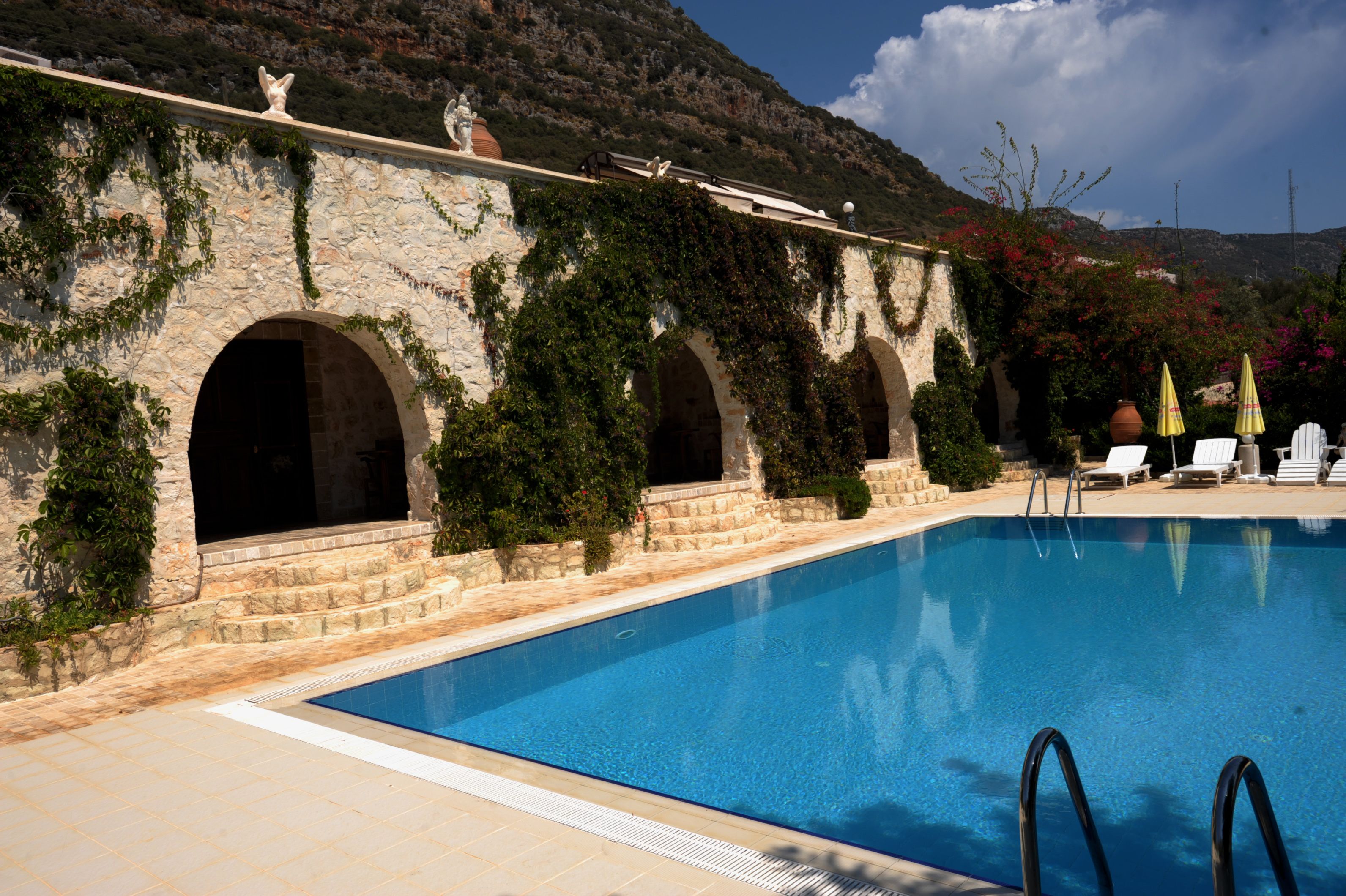 Una piscina con arcos de piedra y vegetación, rodeada de sillas de descanso y sombrillas.