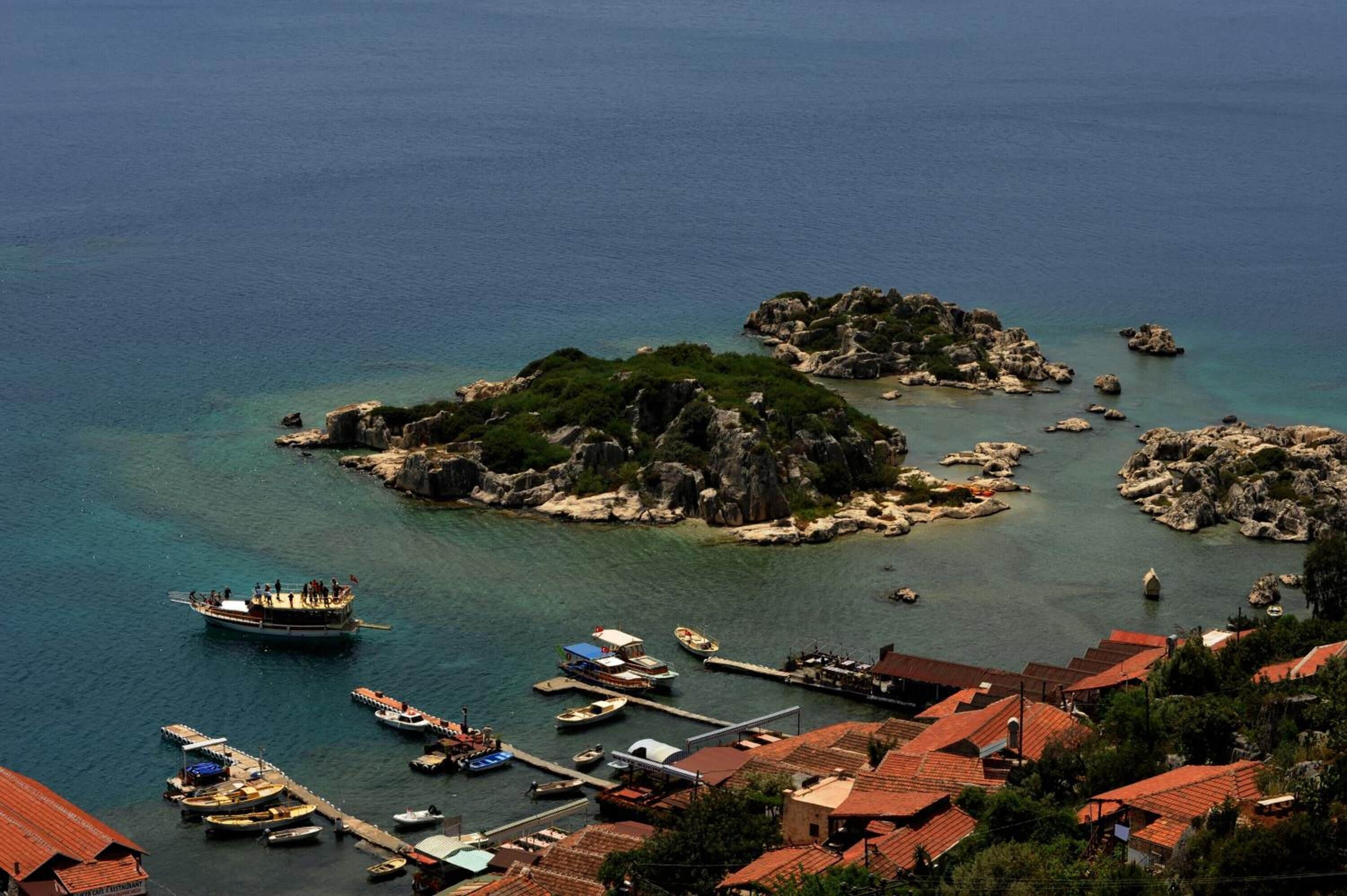 Un pueblo costero con islas rocosas, un muelle con barcos y edificios tradicionales con tejados rojos.