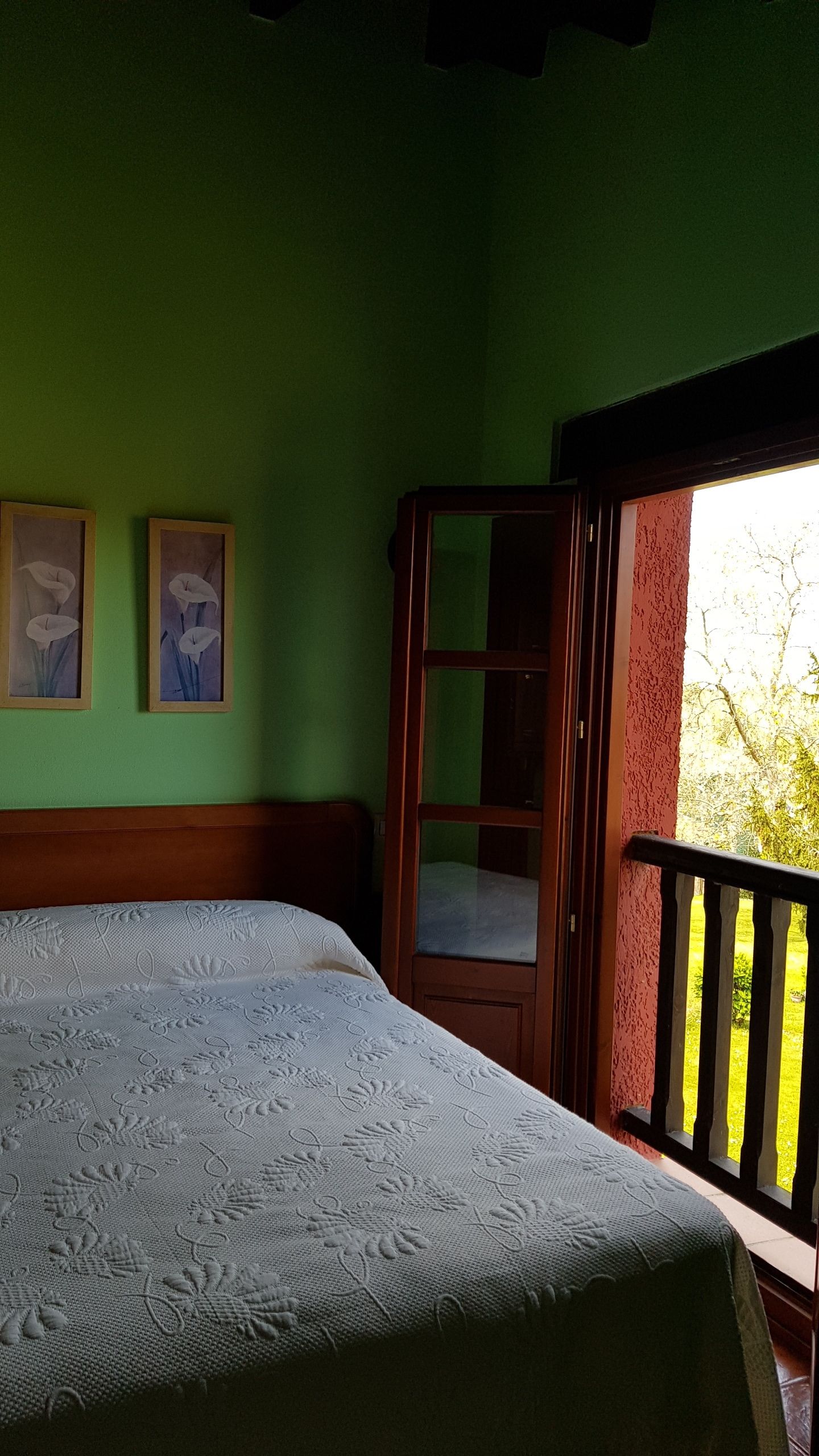Ein Schlafzimmer mit Bett, grünen Wänden, zwei gerahmten Bildern, einem Holzschrank und einem Balkon mit Blick auf Grünflächen.