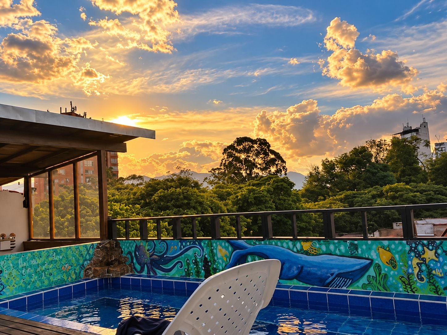 A rooftop pool with a mural, surrounded by trees and buildings, under a sunset sky.