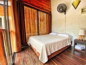 Un dormitorio con una cama de madera, un ventilador de pie y una mesita de noche con una lámpara.