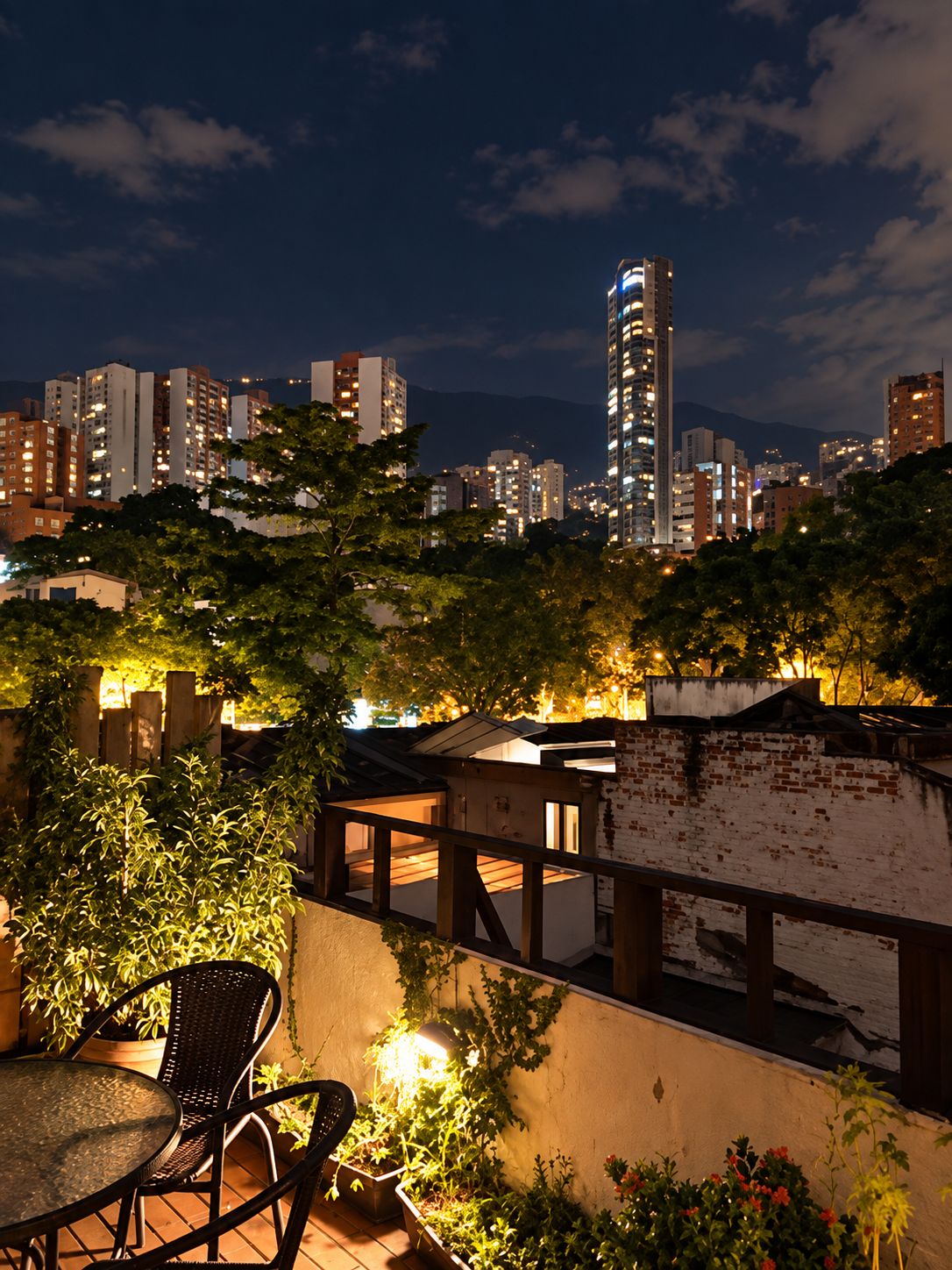 A rooftop terrace with a view of a city skyline at night, including high-rise buildings and a prominent tower.