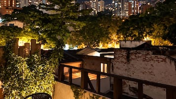 A rooftop terrace with a view of a city skyline at night, including high-rise buildings and a prominent tower.