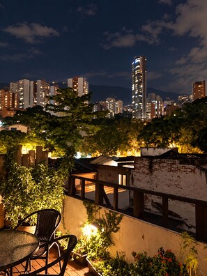 Una terraza en la azotea con vistas a un horizonte urbano nocturno, que incluye edificios altos y una torre destacada.