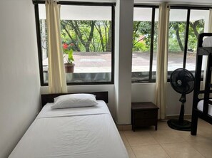 Una cama individual con colchón y almohada blancos, una mesita de noche pequeña, un ventilador de pie y una ventana con cortinas.