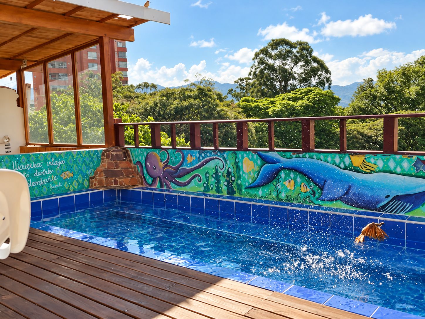 Une piscine ornée d’une fresque représentant la vie marine, entourée d’une terrasse en bois et offrant une vue sur la verdure et les bâtiments.
