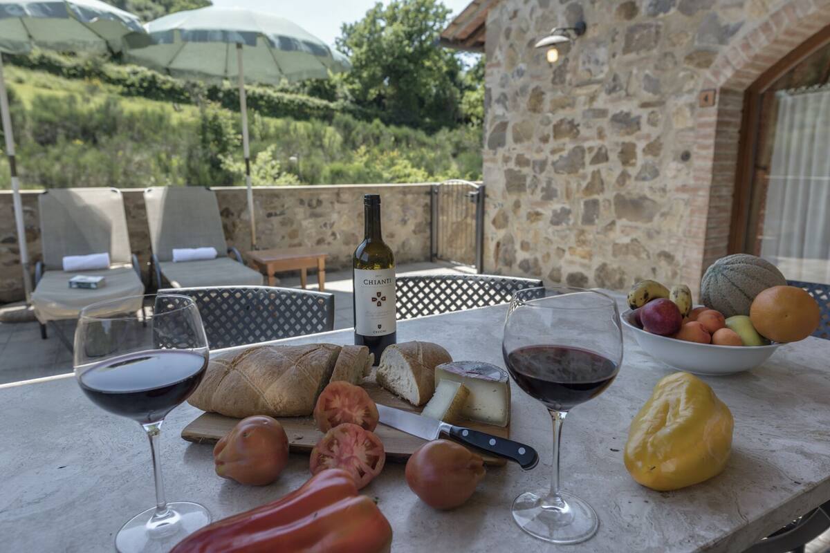 Een tafel gedekt met wijnglazen, kaas, brood en een schaal fruit op een terras met stenen muren en parasols.