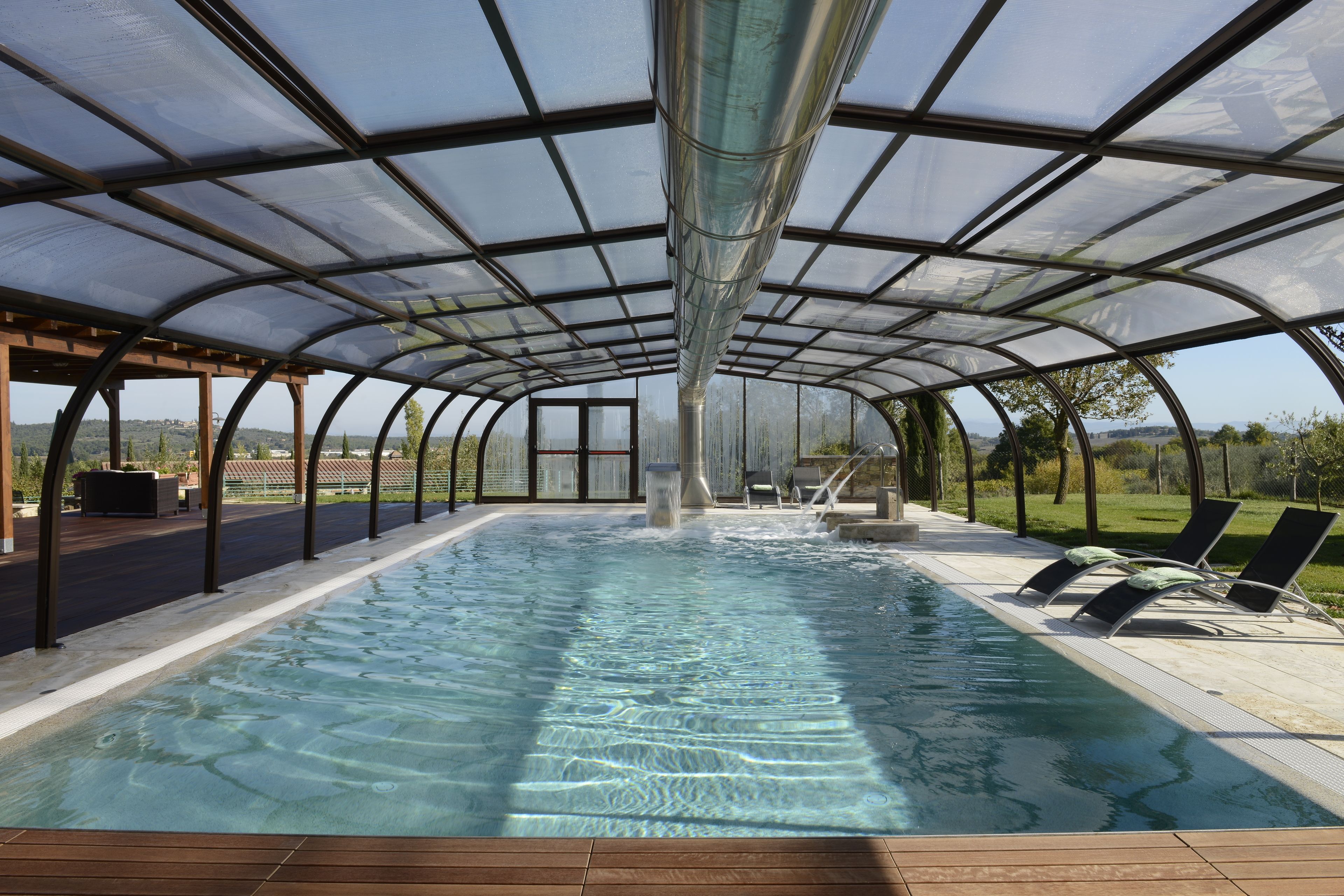 Une piscine intérieure couverte avec un toit en verre, des chaises longues et une vue sur l’extérieur.