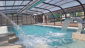 Una piscina interior con techo de cristal, paredes de piedra y un elemento acuático central.