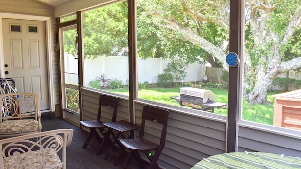 A covered porch with a view of a backyard, featuring a grill, chairs, and a table.