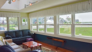 A living room with a dark green couch, a red coffee table, and a patterned rug. There are multiple windows providing a view of a green lawn and a white fence.