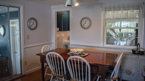 A dining area with a wooden table, white chairs, two wall clocks, a ceiling fan, and a window with a view of trees.