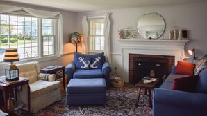 A cozy living room with a blue armchair, a matching ottoman, a fireplace, and a large window with curtains.