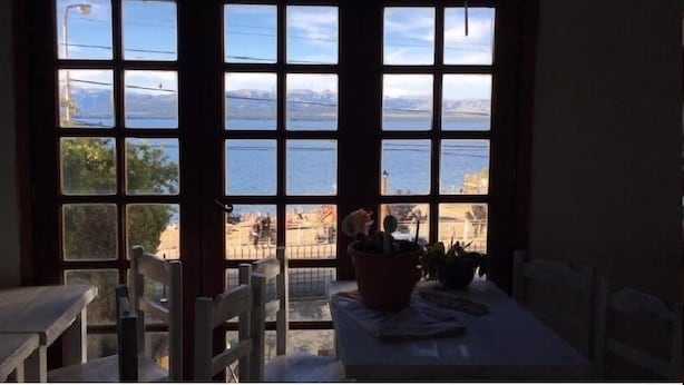 Una habitación con un ventanal grande que da a una masa de agua y un paisaje lejano. Se observa una mesa con una planta en maceta y una silla.