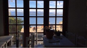 Ein Zimmer mit großem Fenster, das auf ein Gewässer und eine entfernte Landschaft blickt. Es steht ein Tisch mit einem Blumentopf und ein Stuhl im Raum.