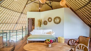 Una habitación con una cama, una mesa de comedor con frutero, una silla de mimbre y vistas al exterior a través de un ventanal.