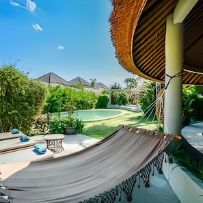 A hammock in a backyard with a pool, a thatched roof structure, and a view of a pyramid-shaped building in the distance.