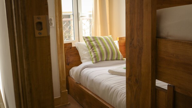 A wooden bed with white bedding and a striped pillow, a window with metal bars, and a wooden door frame.