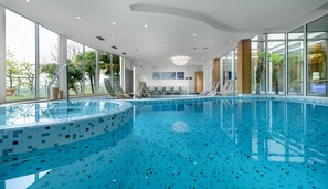 A modern indoor swimming pool with clear blue water, large windows, and sun loungers. - Hotel Wilma (Nalles)