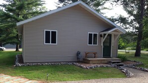 A small, single-story cabin with a front porch, a small deck, and a gravel driveway. - Open All Year. Ausable River Access. Newly Remodeled! (Roscommon)