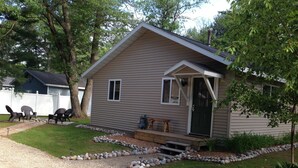A single-story cabin with a front porch, a gravel driveway, and a small garden area. - Open All Year. Ausable River Access. Newly Remodeled! (Roscommon)